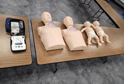 CPR training equipment on tables, including adult torsos, infant mannequins, and a defibrillator.