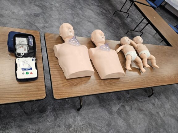 CPR training equipment on tables, including adult torsos, infant mannequins, and a defibrillator.