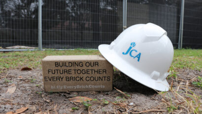 A JCA-branded hard hat and a brick inscribed with "Building Our Future Together Every Brick Counts" placed on the ground.