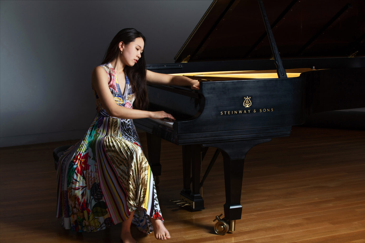 A person in a colorful dress plays a Steinway & Sons grand piano, seated barefoot on a wooden floor.