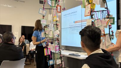 A presentation is taking place in a classroom. A woman stands near a screen displaying a "Values-Based Approach" slide. Hanging notes and decorations are displayed around the screen.