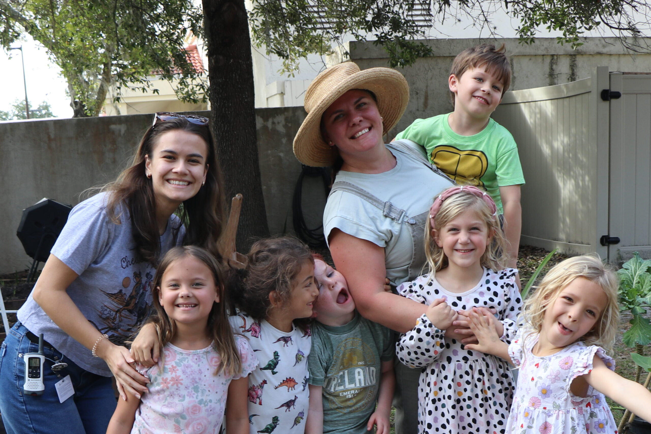 A group of adults and children smiling in a garden, surrounded by plants and raised beds. One adult wears a straw hat.