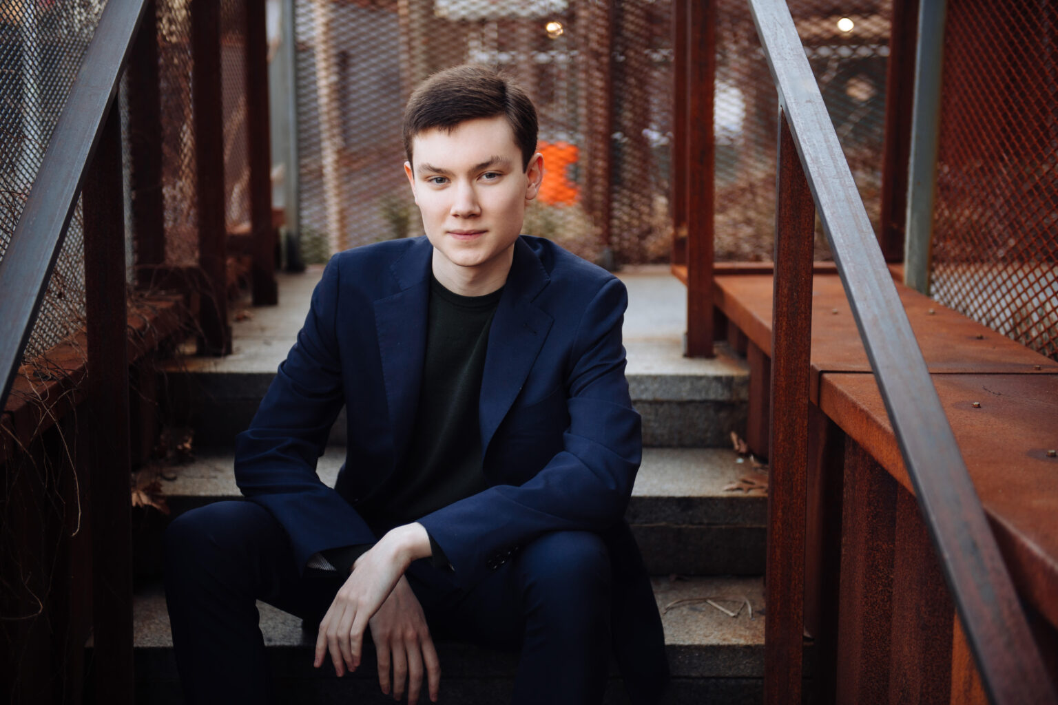 A person in a navy suit sits on outdoor stairs, surrounded by metal railings, looking at the camera.