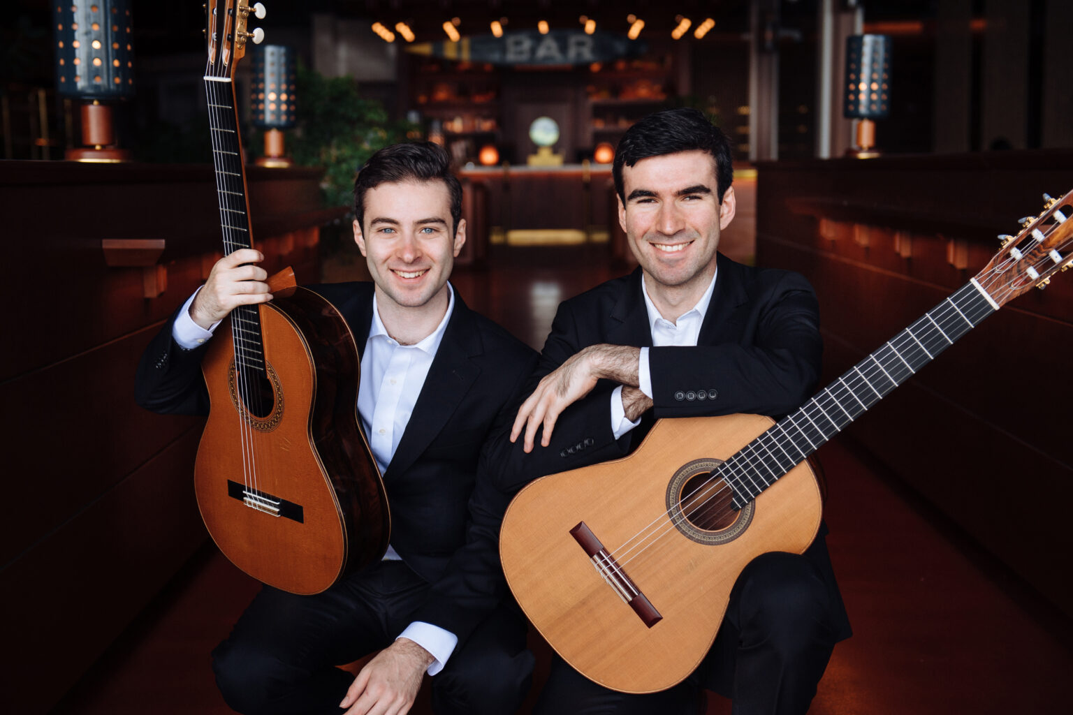 Two men in suits sit side by side, each holding a classical guitar, with a blurred restaurant background.