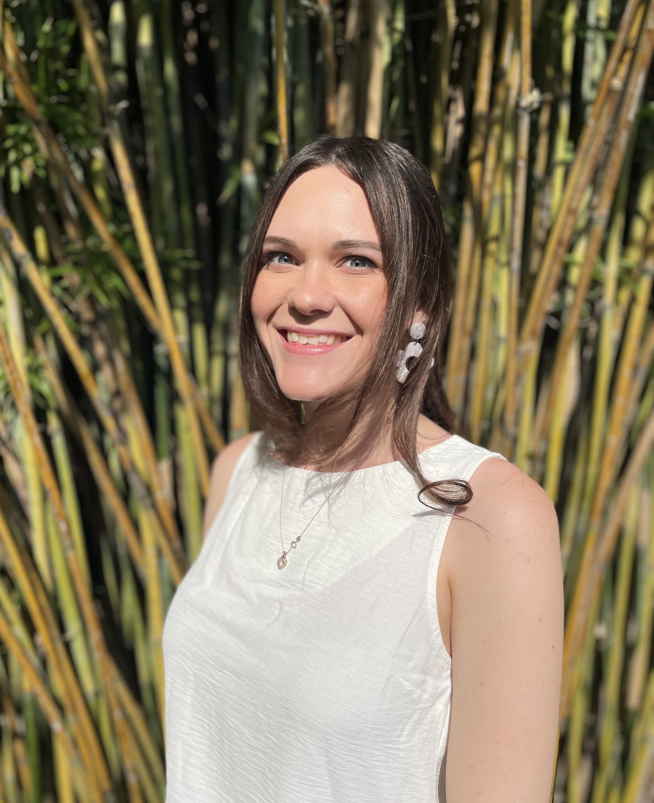 A person with long hair, wearing a white sleeveless top and earrings, is smiling in front of tall bamboo plants.