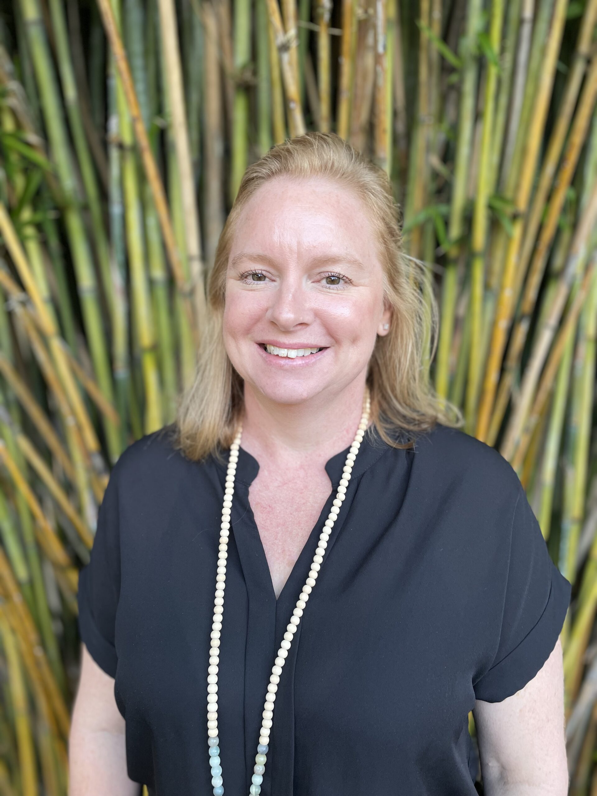 Person with blond hair wearing a black shirt and a beaded necklace, smiling in front of tall bamboo stalks.