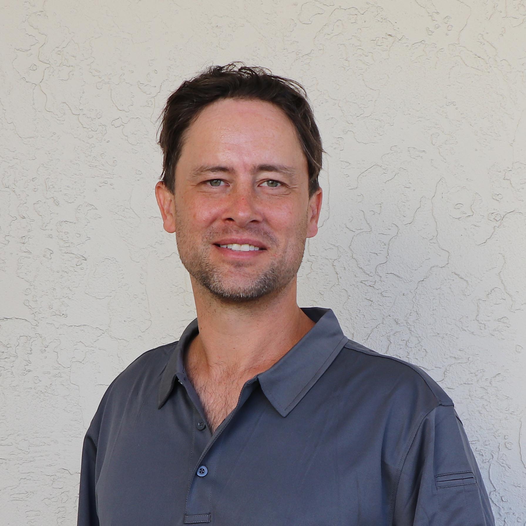 A person with short dark hair and light stubble is smiling slightly while standing against a textured beige wall, wearing a gray button-up shirt.