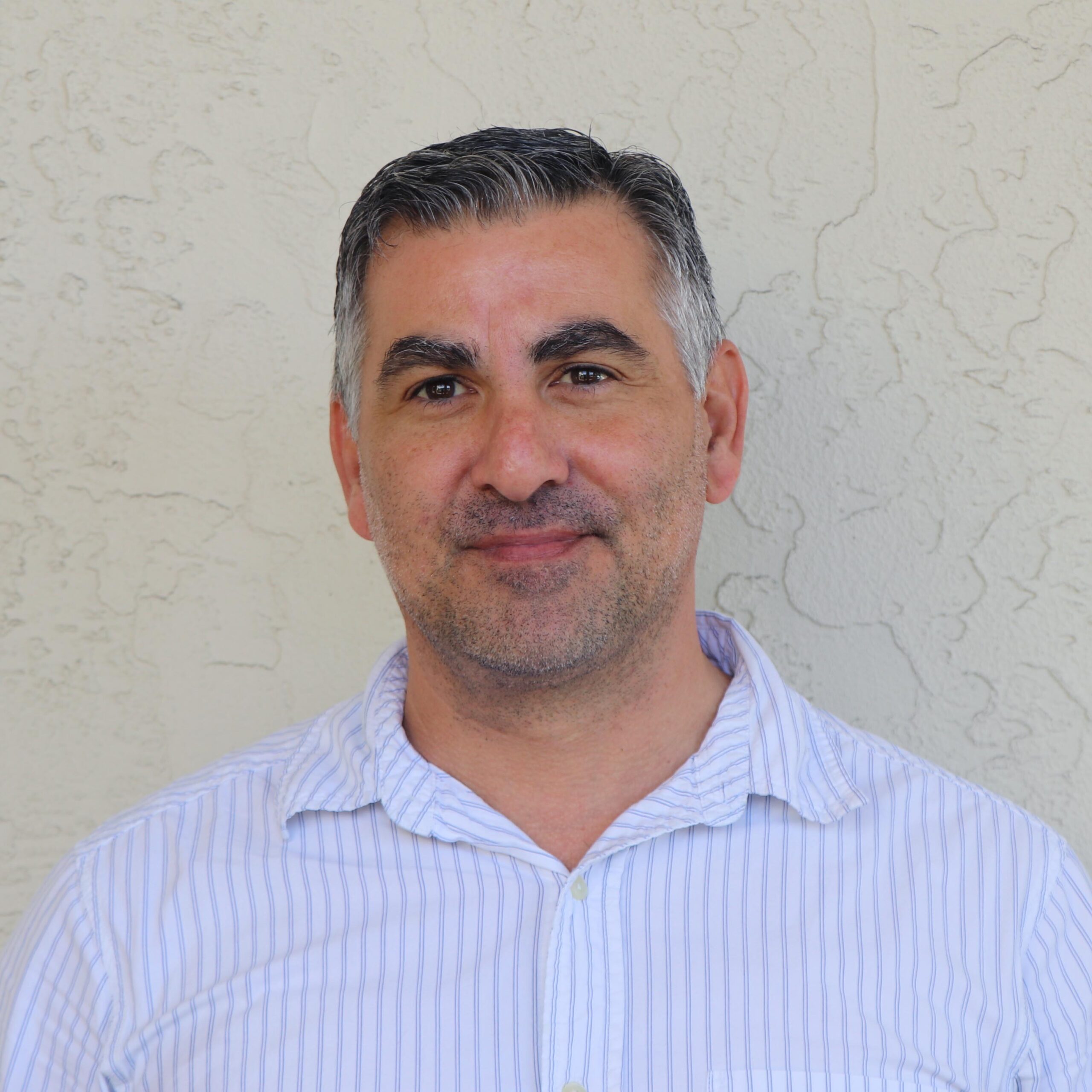 Man with short dark hair and stubble, wearing a light blue striped shirt, standing against a textured beige wall.