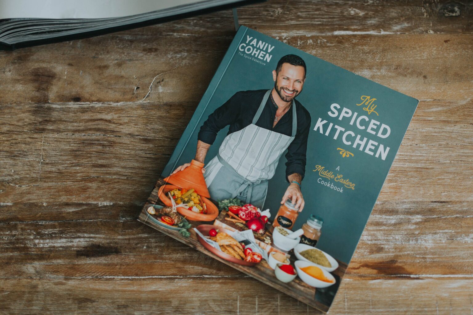 A cookbook titled "My Spiced Kitchen" featuring a man with an apron, surrounded by various spices and dishes, on a wooden table.