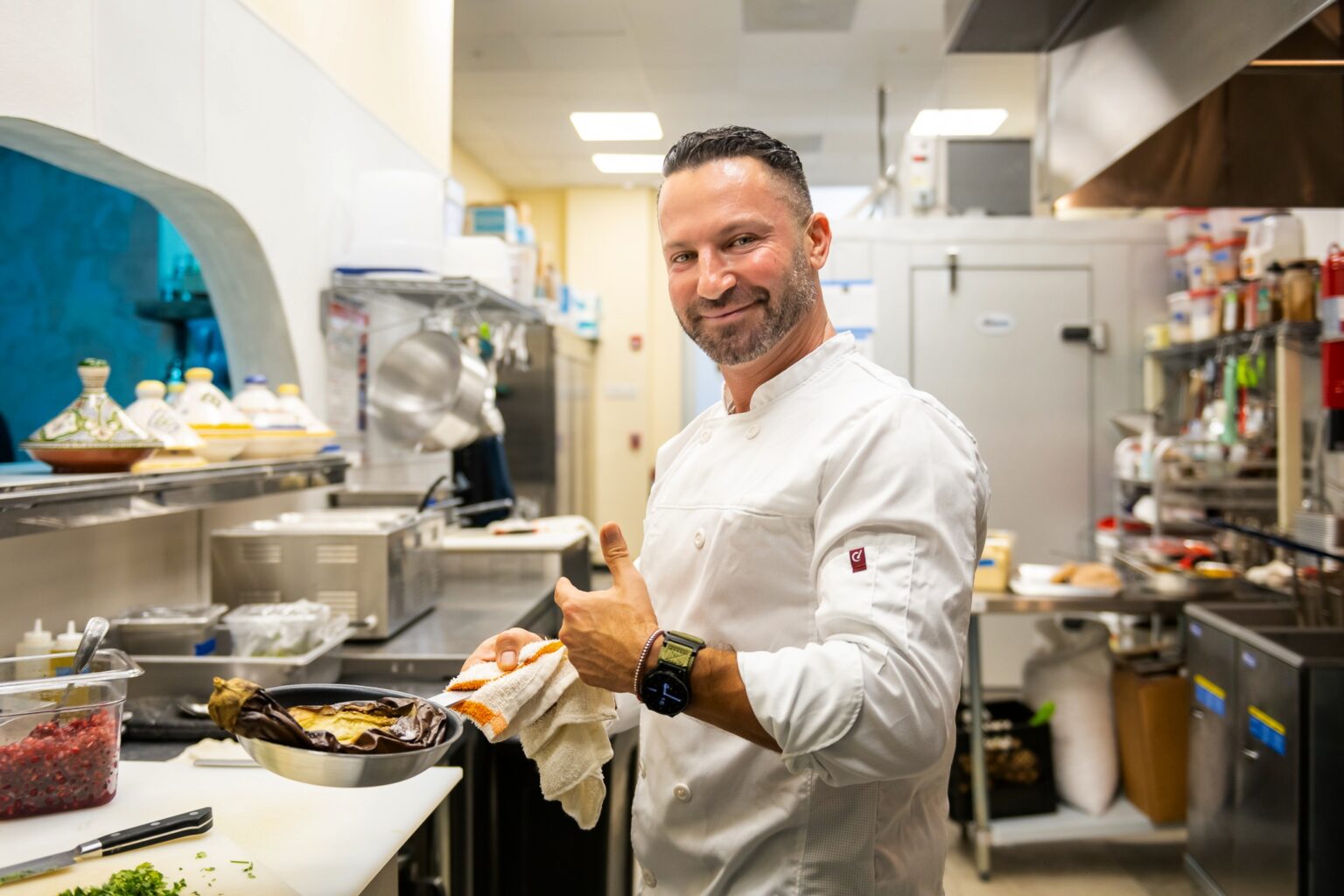 Chef in a white coat gives a thumbs-up in a commercial kitchen, standing beside a counter with ingredients.