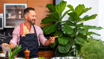 Man in apron gestures while standing in a kitchen surrounded by various vegetables and potted plants.