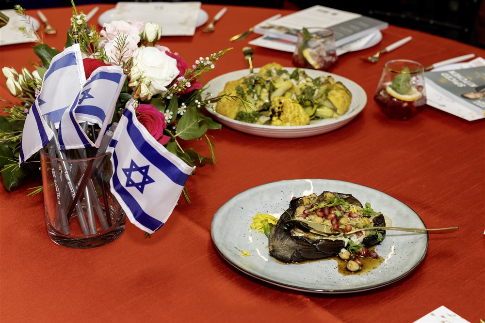 Table setting with a plate of roasted eggplant, a bouquet of flowers with Israeli flags, and a dish of vegetables on a red tablecloth.