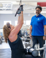 A woman exercises on a lat pulldown machine while a man in a blue shirt stands nearby, observing and smiling in a gym setting.