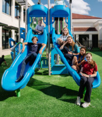 Seven children pose on and around a blue playground slide structure under a green canopy on a sunny day, with school buildings in the background.