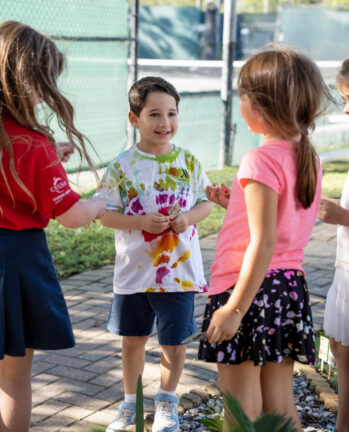 Four children stand and talk together outdoors on a paved path near a tennis court, with trees and greenery in the background.