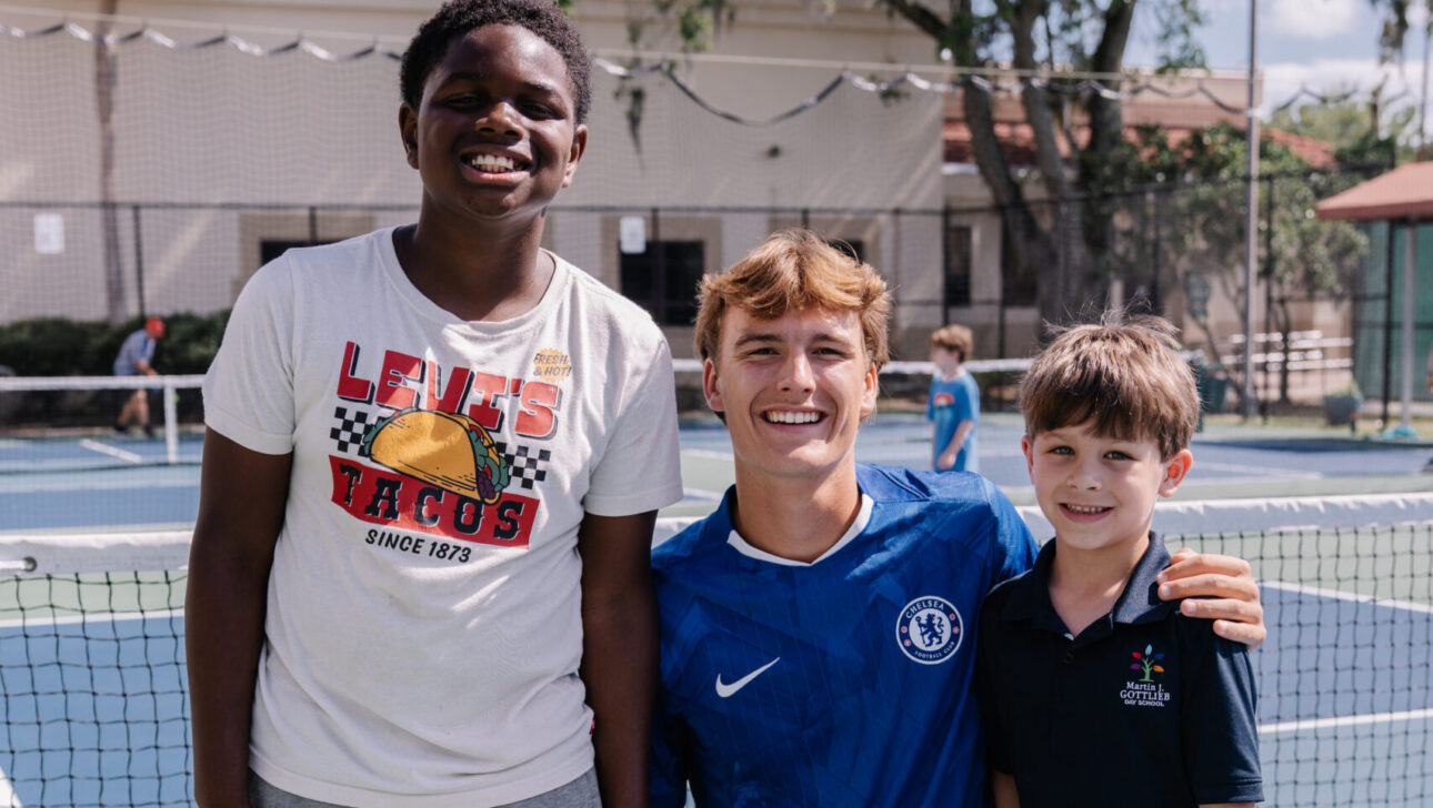 Three boys pose and smile on an outdoor tennis court; one kneels while the other two stand beside him near the net.