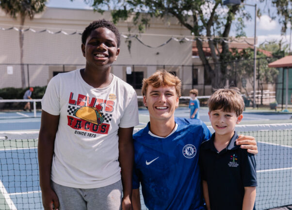 Three boys pose and smile on an outdoor tennis court; one kneels while the other two stand beside him near the net.