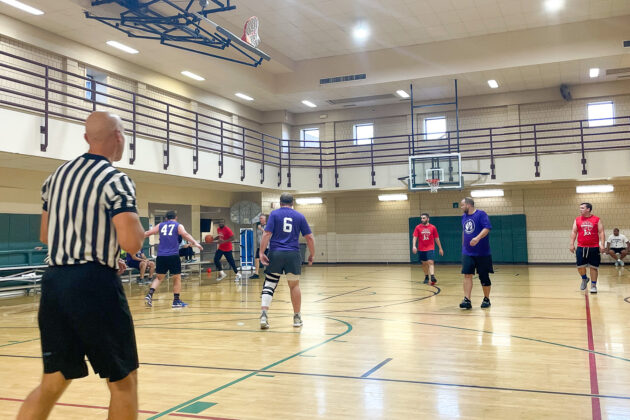 A basketball game is being played in a gym with a referee watching. Players in purple and red jerseys are on the court near the basket.