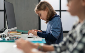 A young girl types on a keyboard at a desk with a computer monitor, while another student works in the foreground.