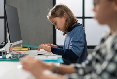 A young girl types on a keyboard at a desk with a computer monitor, while another student works in the foreground.