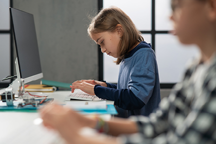 A young girl types on a keyboard at a desk with a computer monitor, while another student works in the foreground.