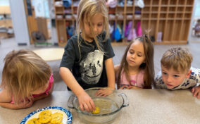 Four young children watch as one dips a round food item into a glass dish, with a plate of similar items on the table in a classroom setting.