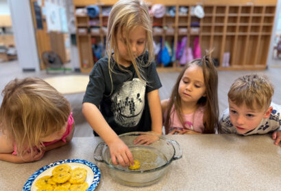 Four young children watch as one dips a round food item into a glass dish, with a plate of similar items on the table in a classroom setting.