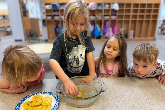 Four young children watch as one dips a round food item into a glass dish, with a plate of similar items on the table in a classroom setting.