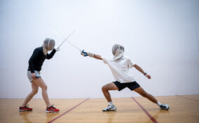 Two people wearing fencing gear spar with swords in a gymnasium, one lunging forward with an extended arm while the other blocks.