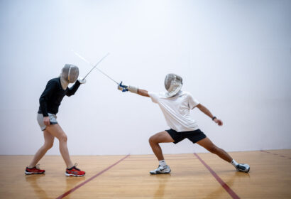 Two people wearing fencing gear spar with swords in a gymnasium, one lunging forward with an extended arm while the other blocks.