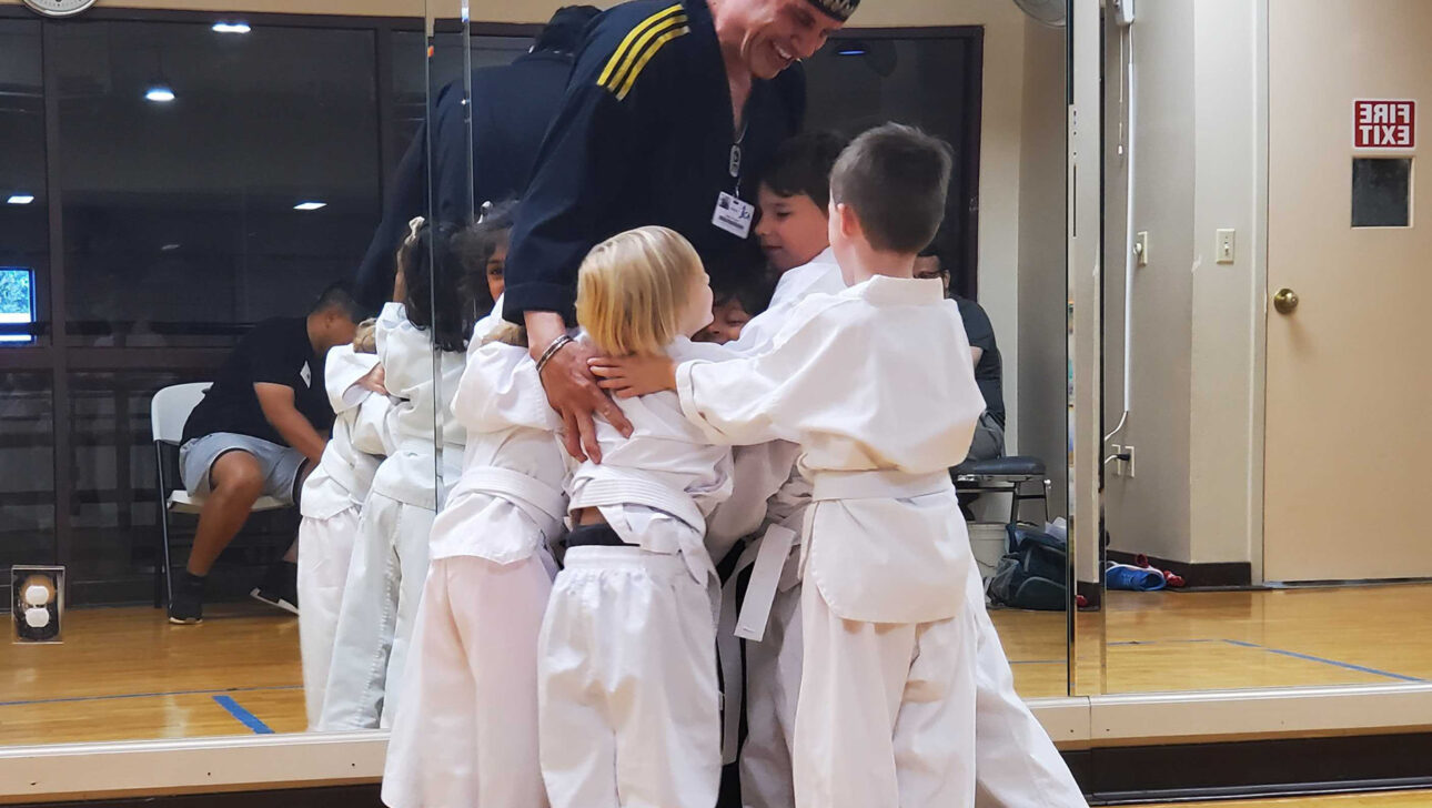 A karate instructor in uniform is standing in a dojo, surrounded by and hugging several young children in white karate uniforms.