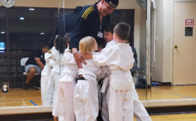 A karate instructor in uniform is standing in a dojo, surrounded by and hugging several young children in white karate uniforms.