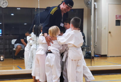 A karate instructor in uniform is standing in a dojo, surrounded by and hugging several young children in white karate uniforms.