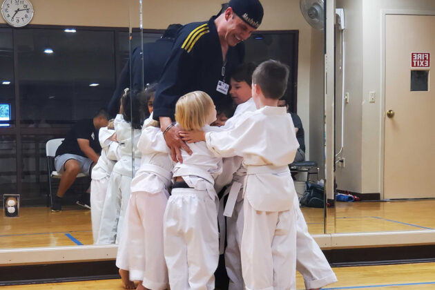 A karate instructor in uniform is standing in a dojo, surrounded by and hugging several young children in white karate uniforms.