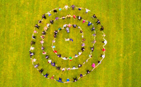 A group of people standing and holding hands in three concentric circles on a grassy field, viewed from above.