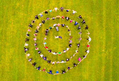 A group of people standing and holding hands in three concentric circles on a grassy field, viewed from above.
