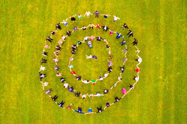 A group of people standing and holding hands in three concentric circles on a grassy field, viewed from above.