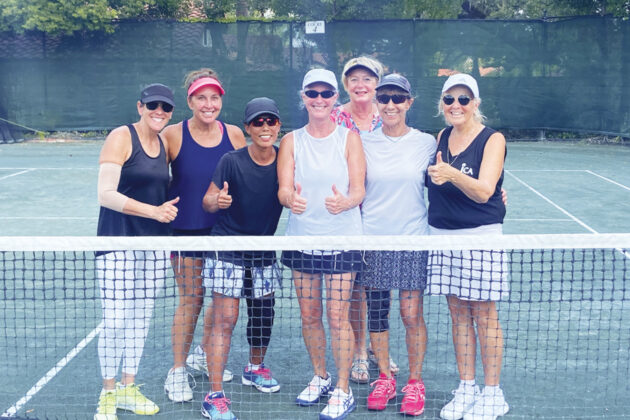 Seven women stand together on a tennis court behind the net, smiling and giving thumbs up, dressed in athletic wear and sunglasses.
