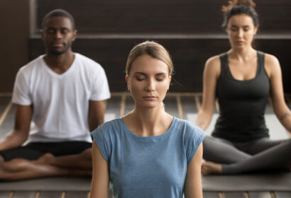 Three people sit cross-legged on yoga mats with eyes closed, meditating indoors.