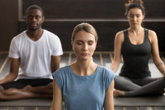 Three people sit cross-legged on yoga mats with eyes closed, meditating indoors.