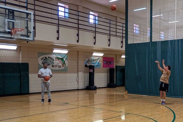 Two people are in a gym; one is shooting a basketball towards the hoop while the other stands holding another basketball, watching.