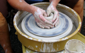 Person shaping clay on a spinning pottery wheel, with hands covered in wet clay; jar and splash marks visible nearby.