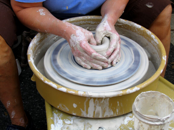 Person shaping clay on a spinning pottery wheel, with hands covered in wet clay; jar and splash marks visible nearby.