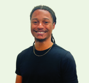 A man with long braids, wearing a black shirt and a gold necklace, smiles at the camera against a plain light background.