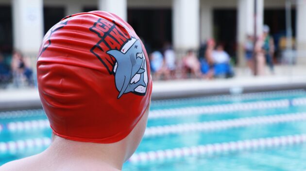 A boy wearing a red swim cap in front of a swimming pool.