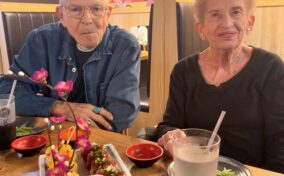 An elderly man and woman sit at a restaurant table with drinks and sushi dishes, against a vibrant, colorful mural background.