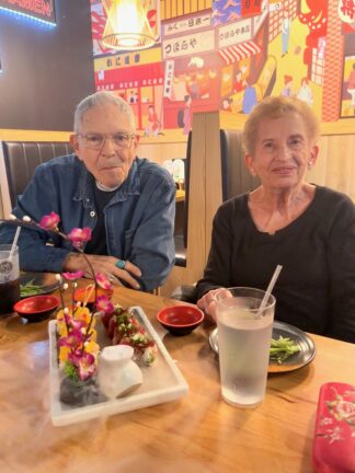 An elderly man and woman sit at a restaurant table with drinks and sushi dishes, against a vibrant, colorful mural background.