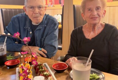 An elderly man and woman sit at a restaurant table with drinks and sushi dishes, against a vibrant, colorful mural background.