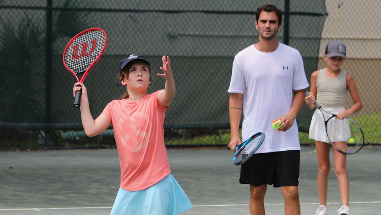 A young person tosses a tennis ball to serve on an outdoor court, while an adult and another child watch nearby, holding rackets and tennis balls.