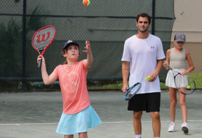 A young person tosses a tennis ball to serve on an outdoor court, while an adult and another child watch nearby, holding rackets and tennis balls.